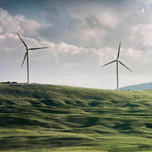 Δύο ανεμογεννήτριες στην κορυφή ενός πράσινου λόφου, με σύννεφα και λιακάδα στο βάθος.   Δύο ανεμογεννήτριες στην κορυφή ενός πράσινου λόφου, με σύννεφα και λιακάδα στο βάθος.